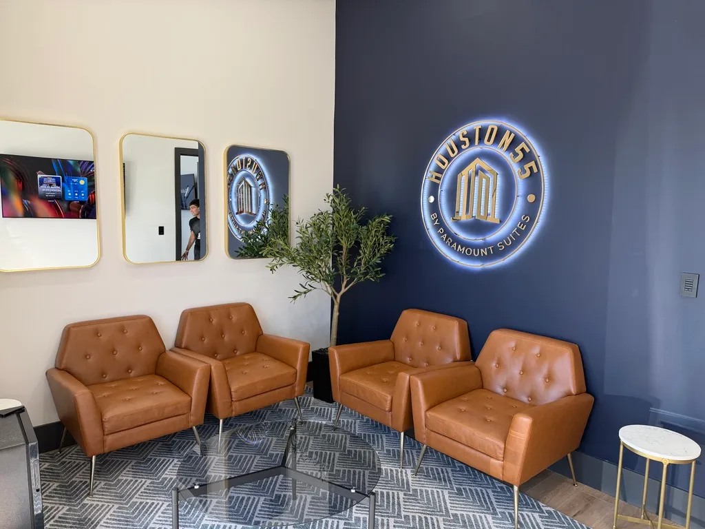 Interior lounge of Houston55 by Paramount Suites, with tan leather chairs, gold-framed mirrors, and the illuminated Houston55 emblem on a navy accent wall.